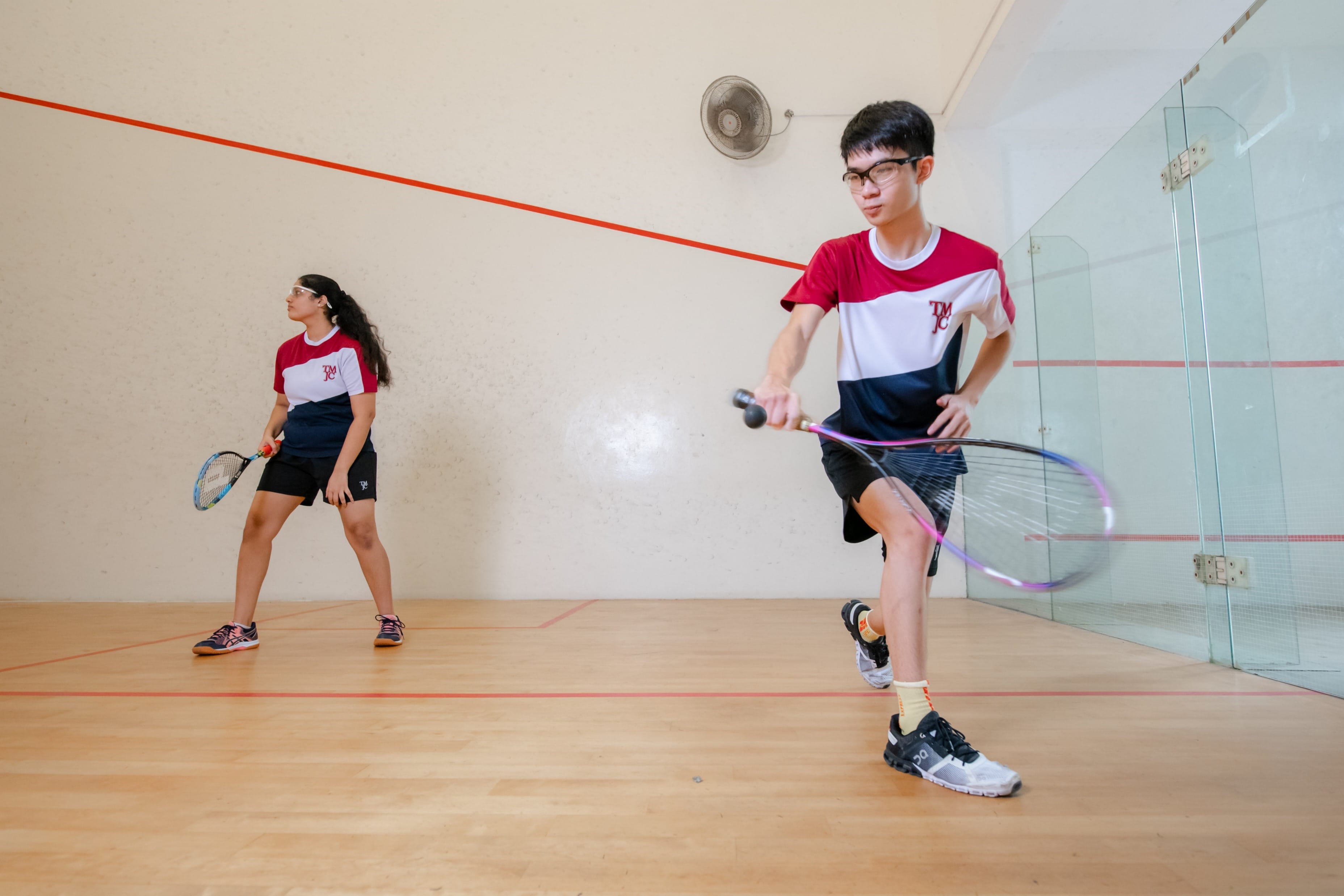 Two young squash players in matching red and white uniforms playing on an indoor squash court with glass walls.